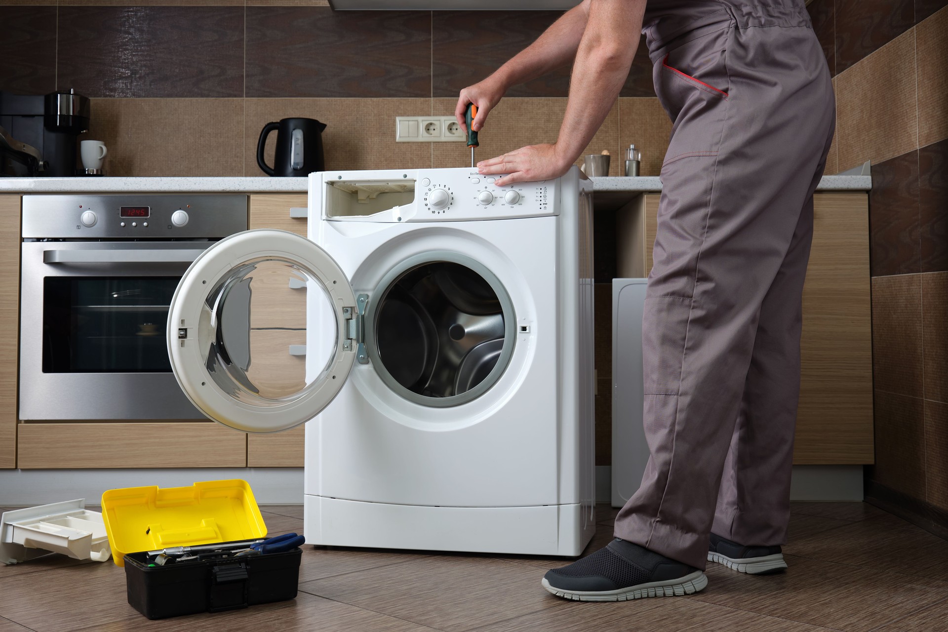 A adult household appliance repairman disassembling a washing machine with a screwdriver.
